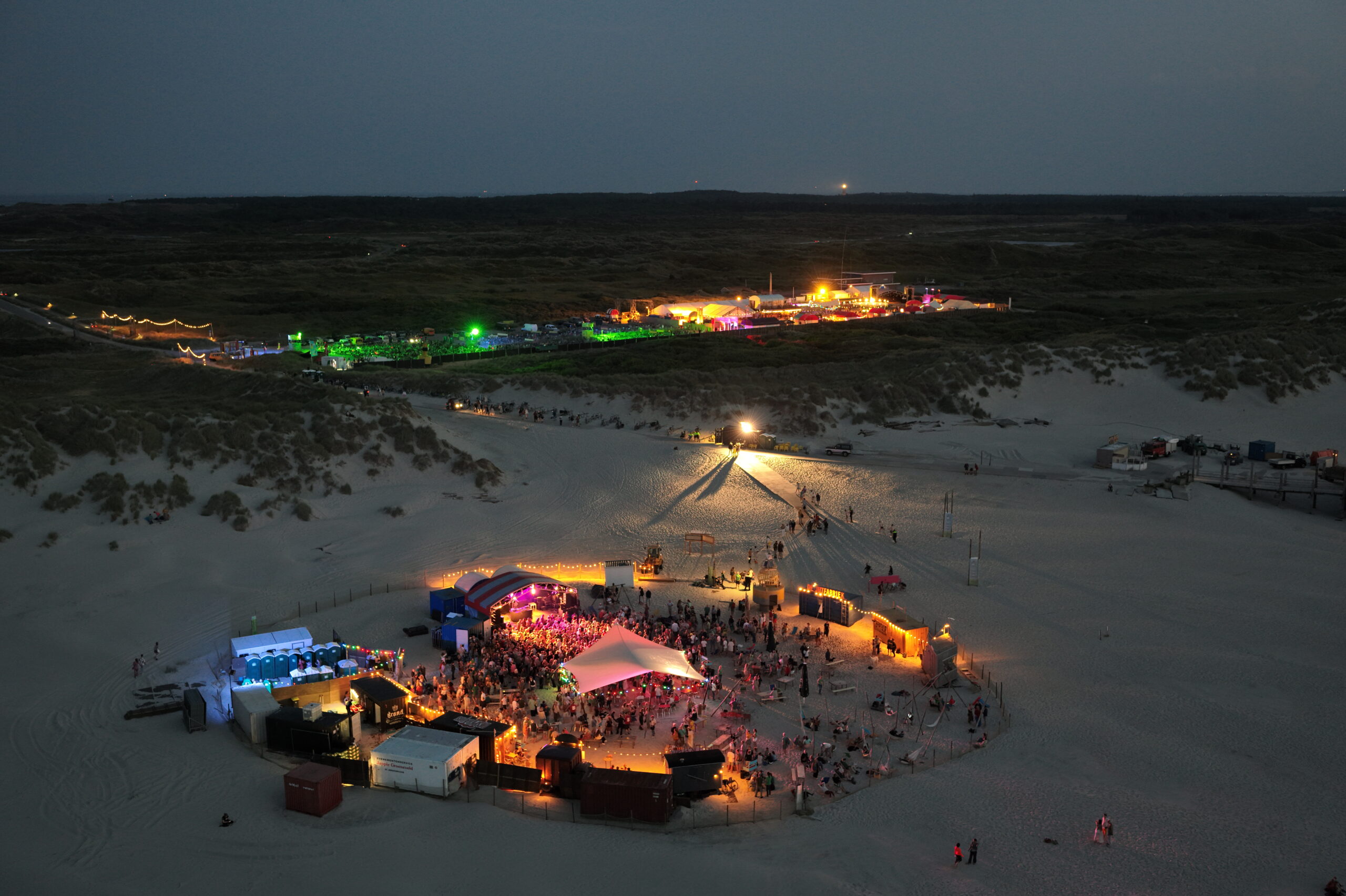 Droneshot van de sfeervol verlichte festivalterreinen het Strand en de Deining op een avond tijdens Oerol 2023.
