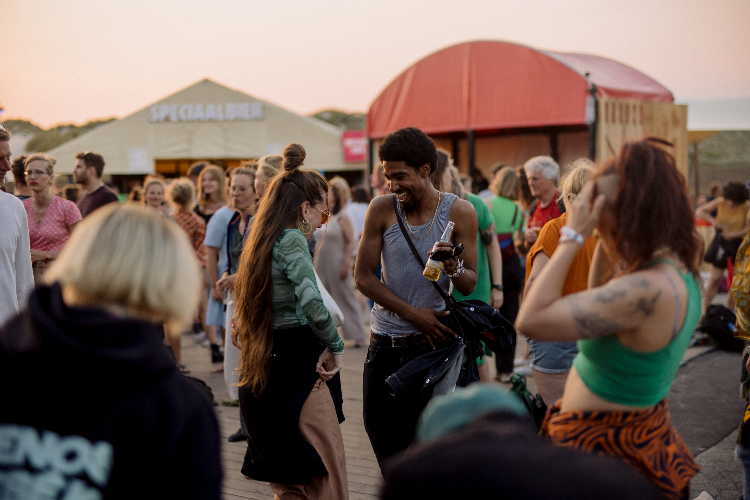 Twee jonge mensen dansen en lachen op festivalhart de Deining tijdens zonsondergang, Oerol 2023
