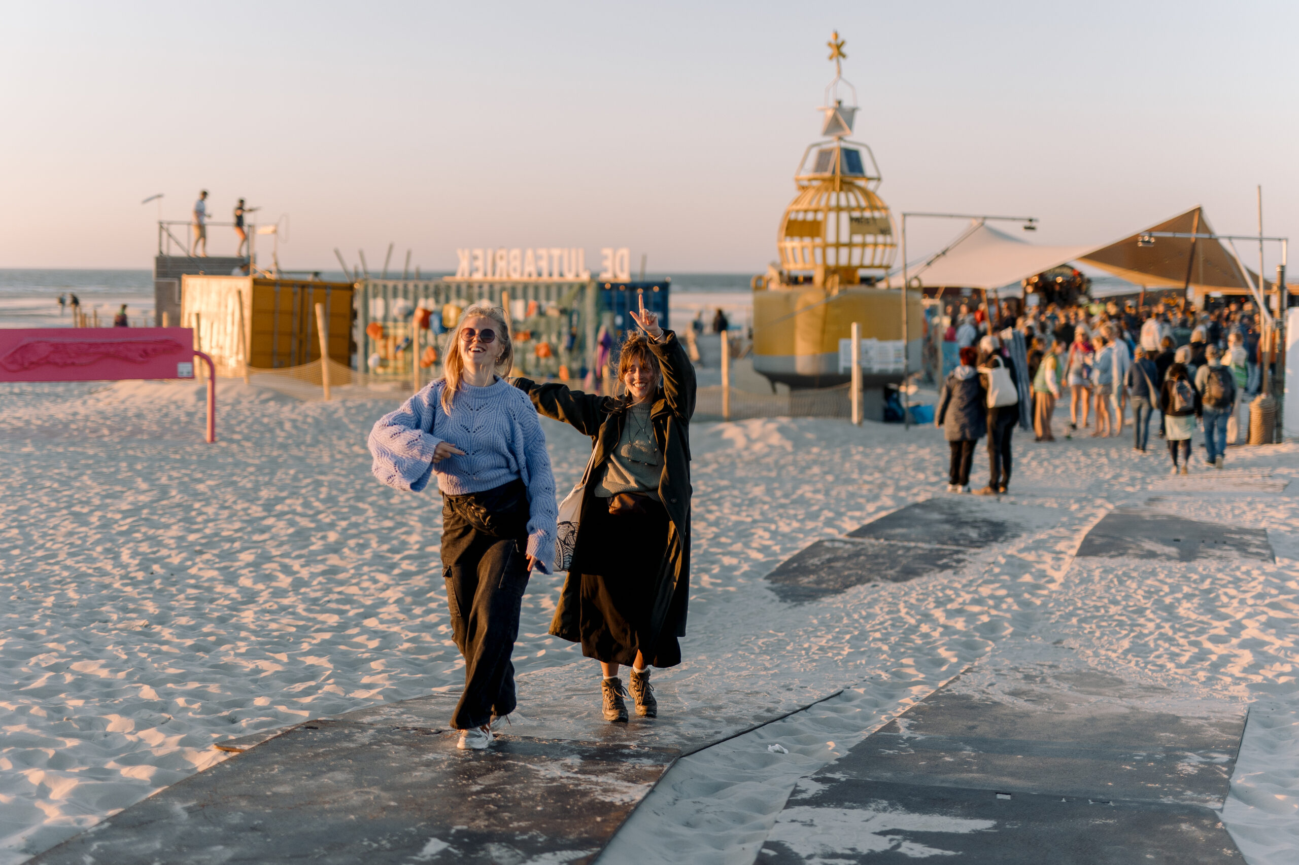 Twee jonge vrolijke vrouwen lopen het festivalhart het Strand af in het licht van de ondergaande zon tijdens Oerol 2023.