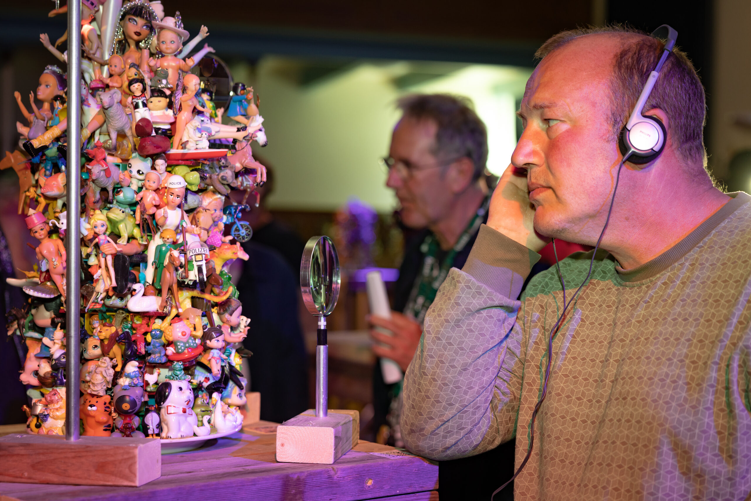 Een man luistert via een koptelefoon naar het verhaal achter een van de Torens van Nienke, een toren van ongeveer 50 centimeter hoog welke volledig bekleed is met speelgoedfiguurtjes, onderdeel van een expositie in de kerk van Midsland op Terschelling tijdens Oerol 2023.