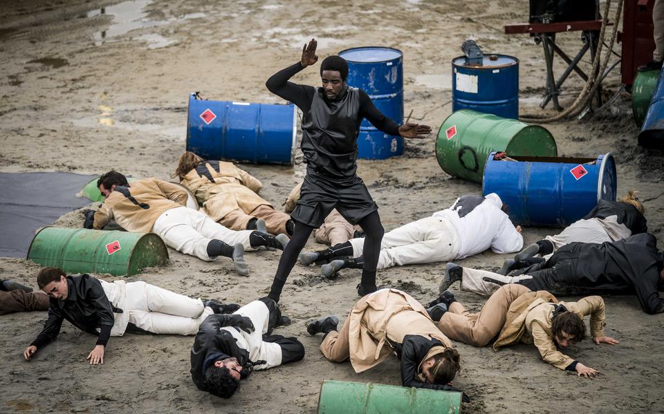 Een zwartgeklede danser staat midden in een cirkel van mensen in wit met zwart besmeurde pakken die op de grond liggen, omringt door oliedrums met rode stickers die waarschuwen voor de giftige inhoud. Een beeld uit de voorstelling Kaapdiegoeiekoop tijdens Oerol 2022.