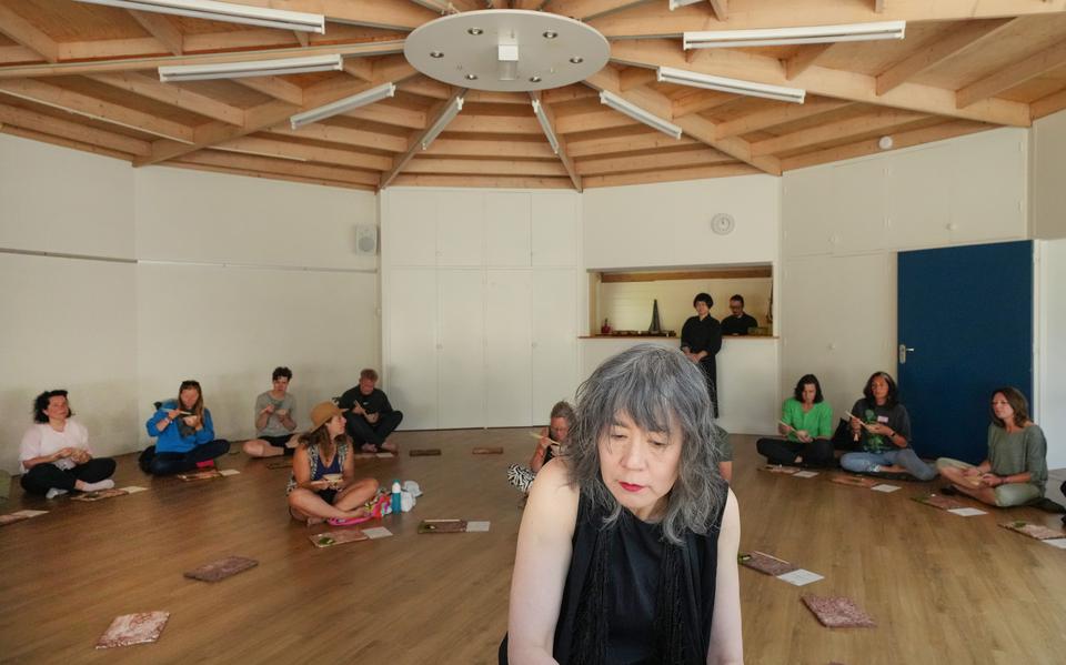 Japanse theatermaker en pianist Tomoko Mukaiyama speelt tijdens Oerol 2023 een compositie op de piano, terwijl het publiek om haar heen op grote afstand van elkaar op de grond zit en met stokjes een ontbijt geserveerd in een klein kommetje nuttigt.