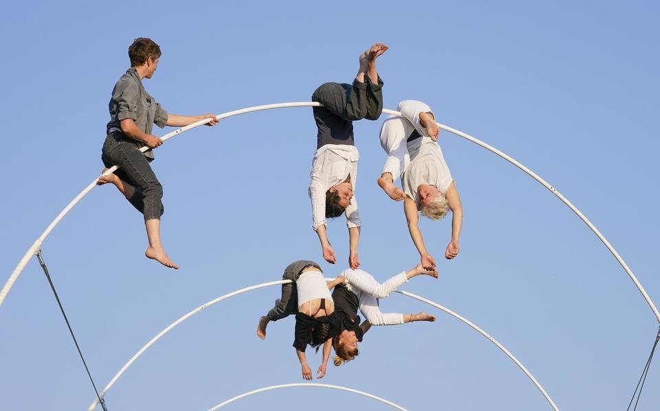 Hoog in de blauwe lucht hangen vier acrobaten ondersteboven in spiraalvormige cirkels die het decor vormen van theatervoorstelling La spire tijdens Oerol 2023.