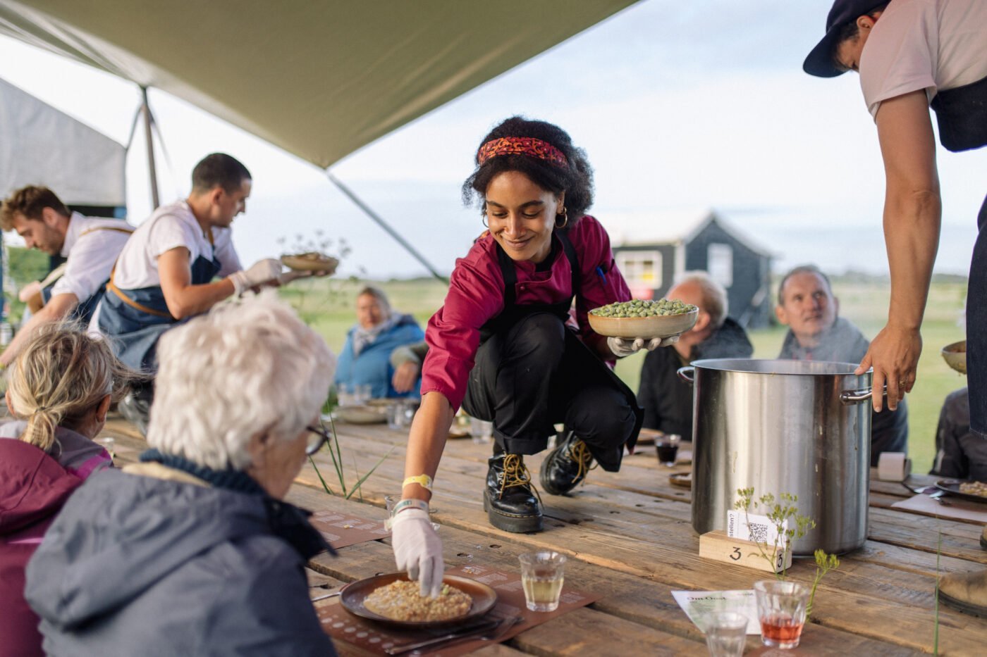 een lange tafel met grote pannen en mensen die eten tijdens Diners om Oost, Marenthe van der Valk, Oerol 2022