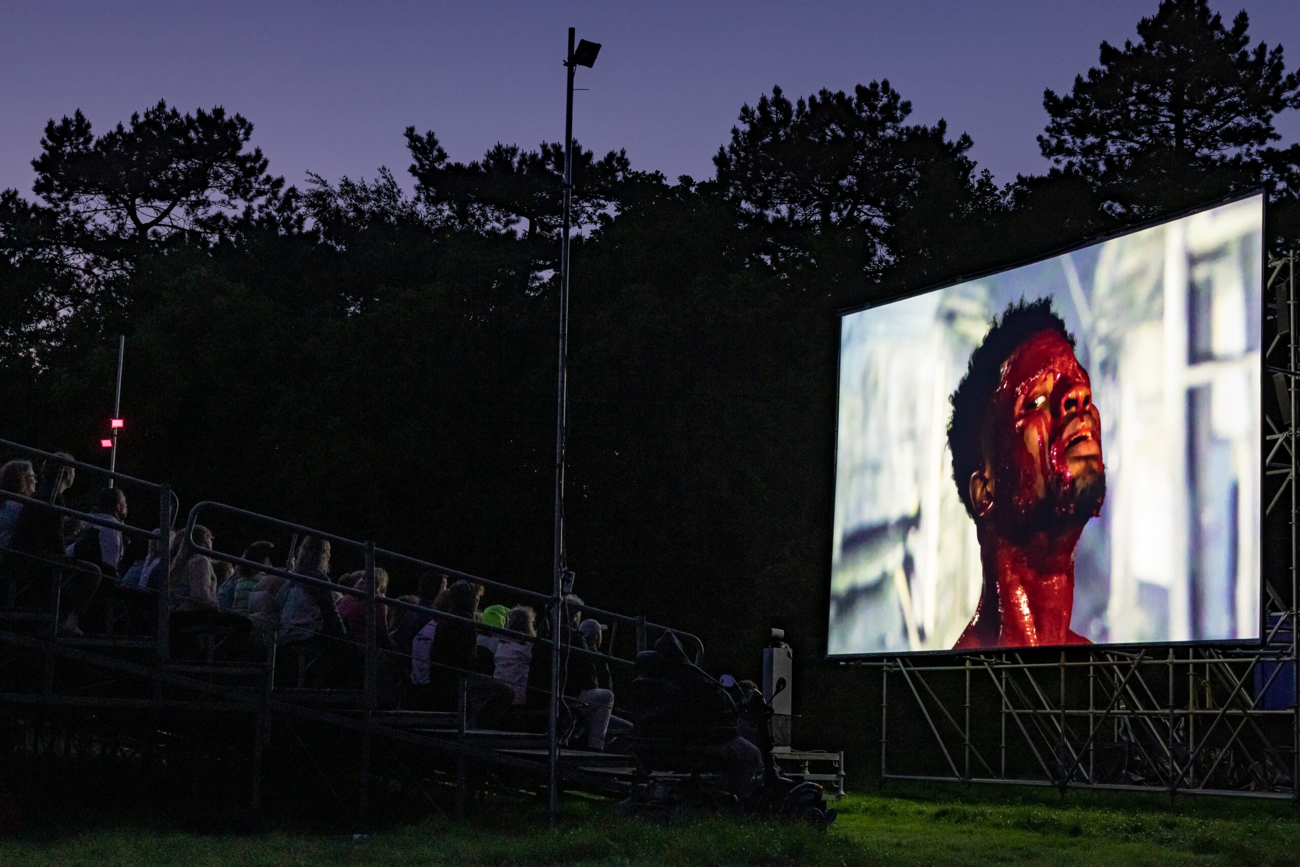 Publiek kijkt naar een groot beeldscherm waar een acteur te zien is via een live-verbinding met Nigeria tijdens de theatervoorstelling Dear beloved Friend van theatermaker Dries Verhoeven tijdens Oerol 2023.