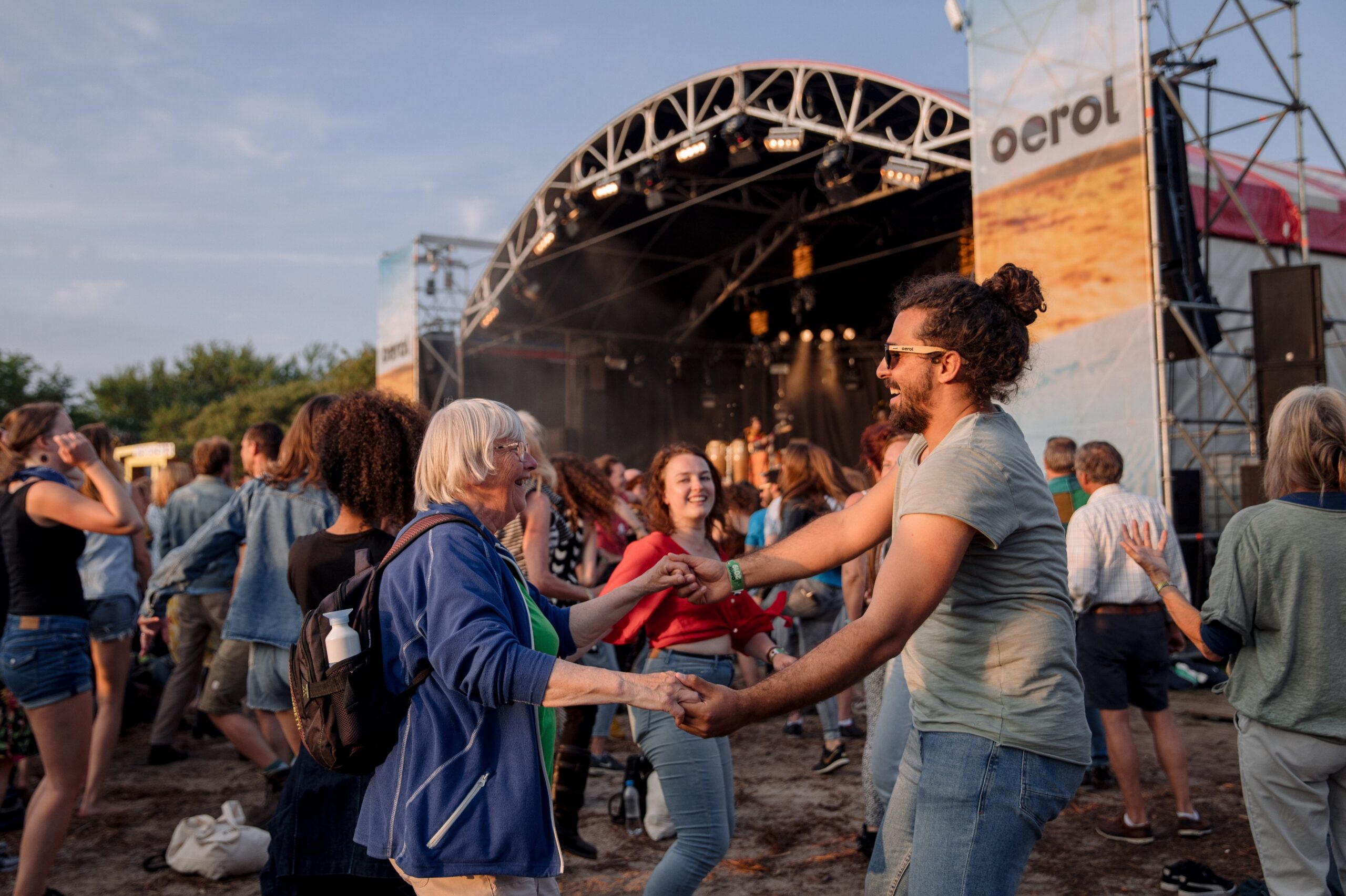 foto van een oude grijze vrouw en een jonge man met knot voor het podium, die hand-in-hand met elkaar dansen met een grote lach op hun gezichten