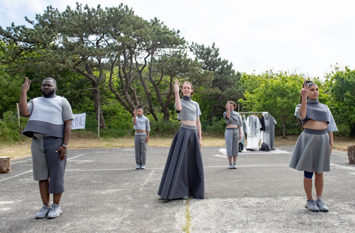 5 acteurs in grijs gekleed steken 3 vingers in de lucht, een voorstellingsbeeld uit Gendermonologen van Raymi Sambo Maakt gespeeld op Oerol 2023