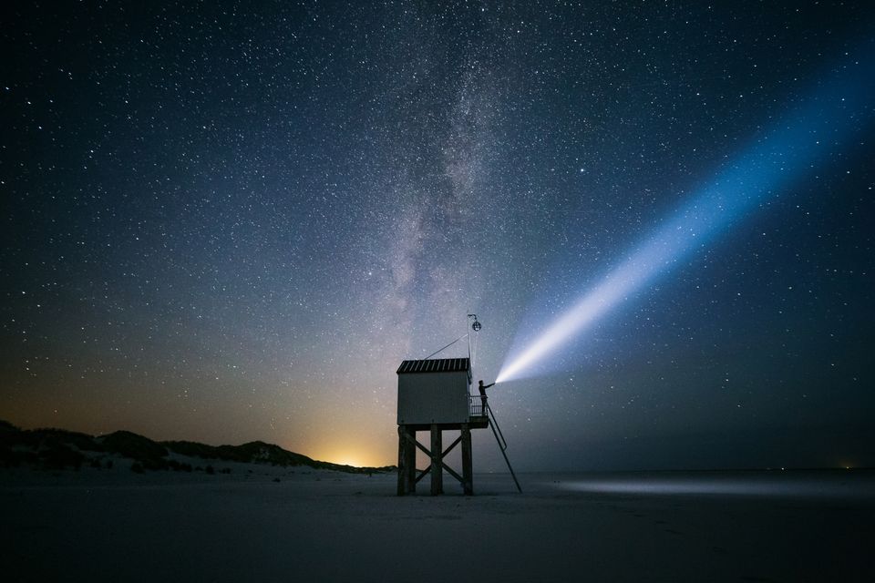 Het drenkelingenhuisje op de Boschplaat op Terschelling onder een donkere sterrenhemel met op de achtergrond het licht van vuurtoren de Brandaris.