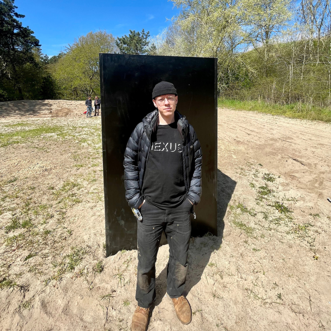 Een persoon in het zwart gekleed staat in het bos voor een zwarte doos die hoger is dan de persoon.