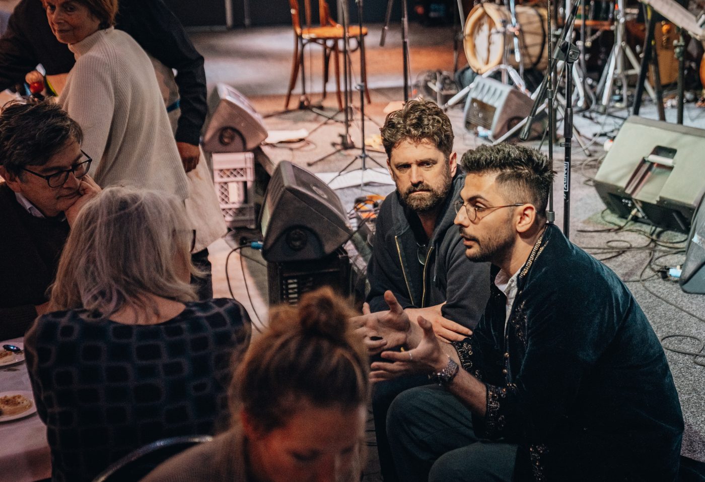 Een groep mensen zit in gesprek in een informele setting, mogelijk tijdens een culturele bijeenkomst. Op de voorgrond spreekt een jongeman met bril en gebaren, terwijl een man naast hem aandachtig luistert. Op de achtergrond staan muziekinstrumenten en geluidsapparatuur op een podium. De sfeer is betrokken en geconcentreerd.