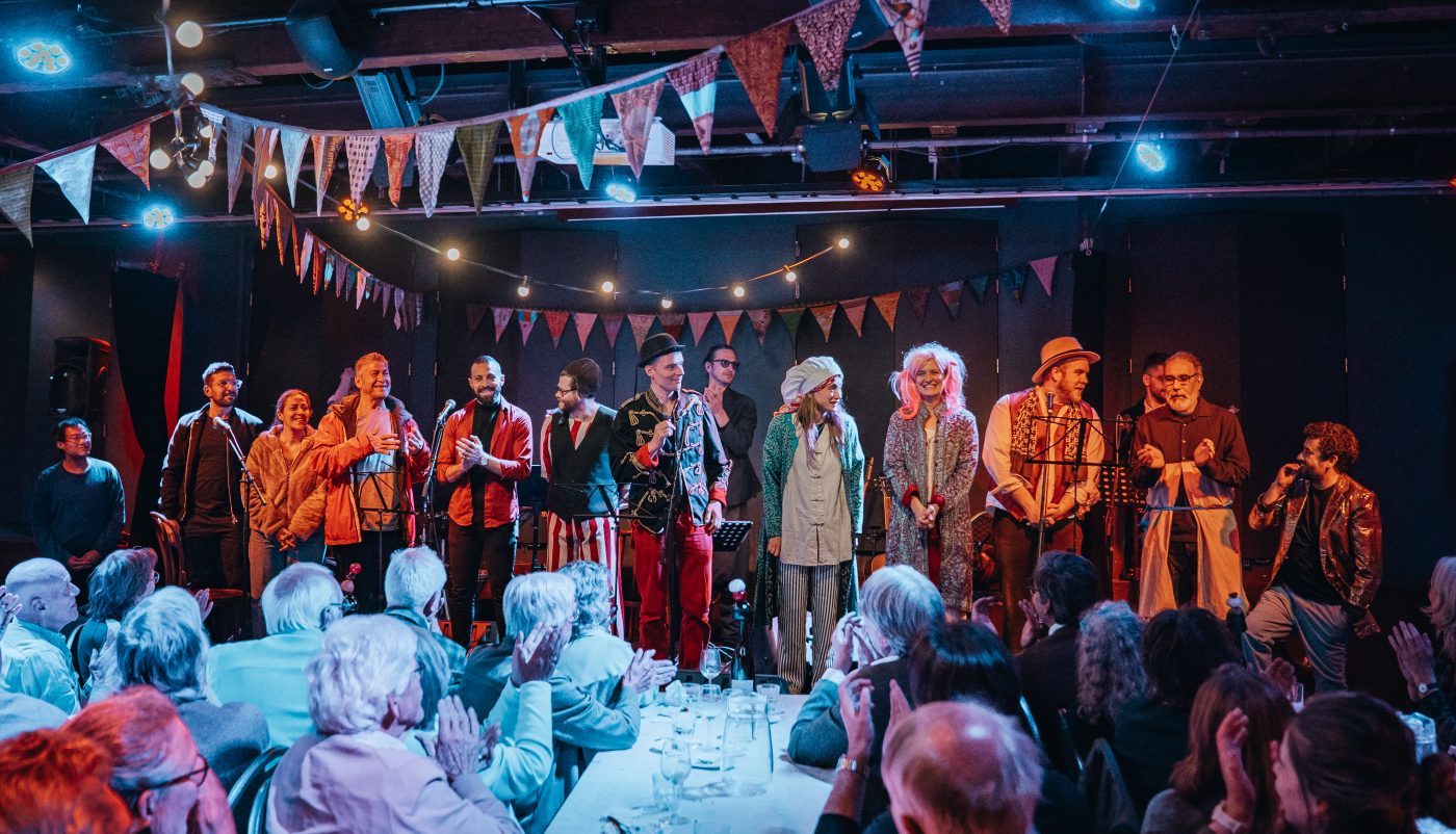 Een groep performers staat op een rij op het podium en ontvangt enthousiast applaus van het publiek.