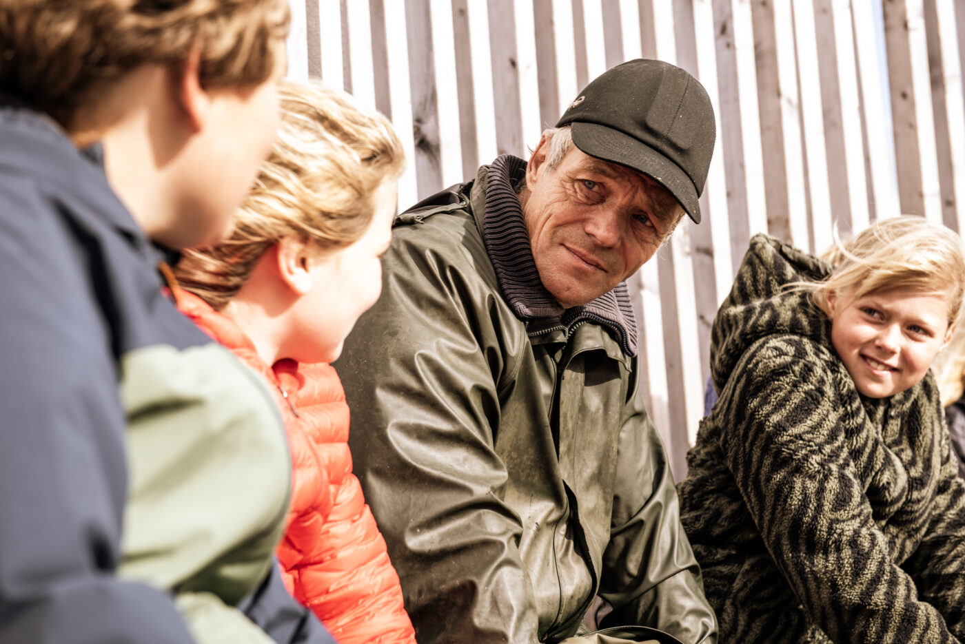 Een oudere man in een groene regenjas en zwarte pet zit buiten op een bankje en praat met drie kinderen die om hem heen zitten. Ze dragen warme jassen en luisteren met aandacht, terwijl de zon op hun gezichten schijnt. Op de achtergrond staan houten palen.