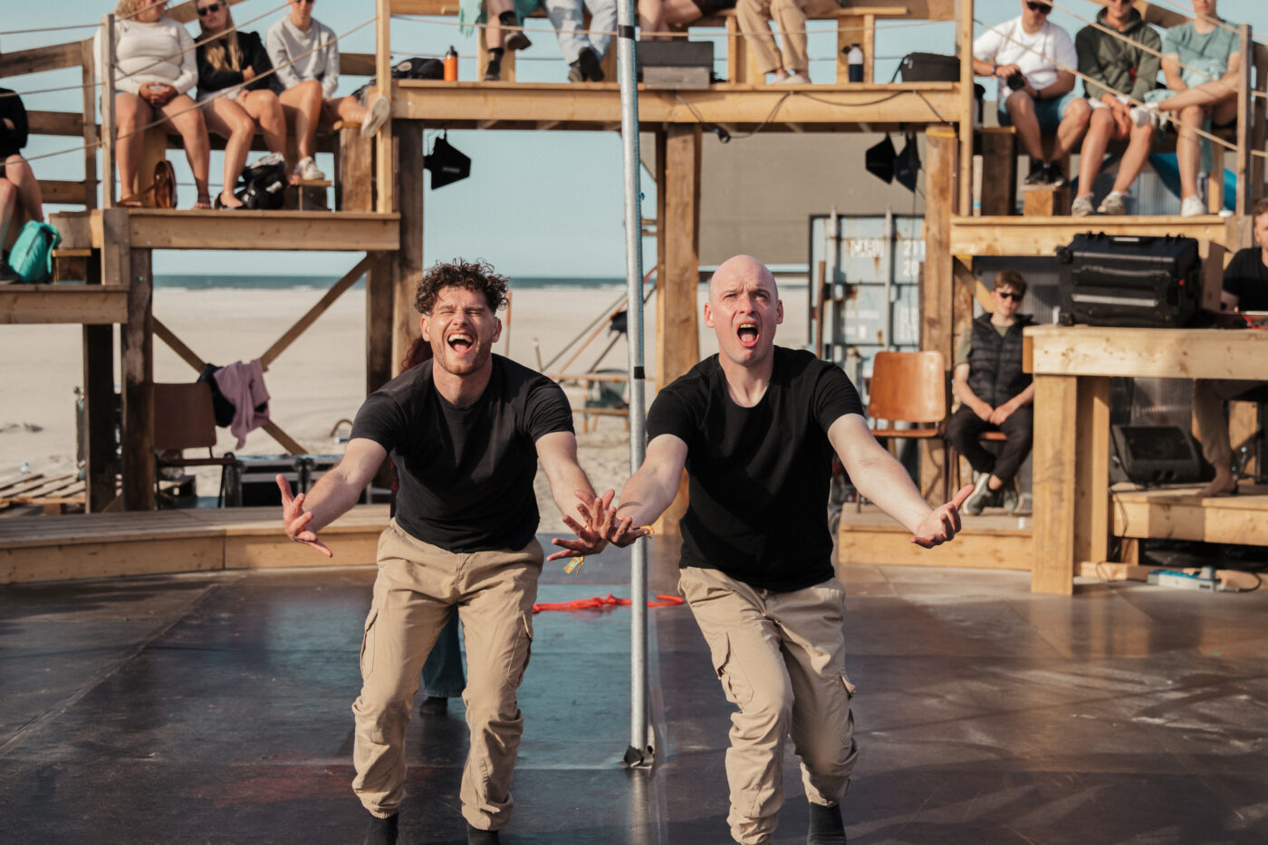 Twee performers in zwarte shirts en beige broeken staan expressief op een buitenpodium, met open mond en uitgestrekte armen naar het publiek. Achter hen zitten toeschouwers op houten tribunes, met uitzicht op strand en zee.
