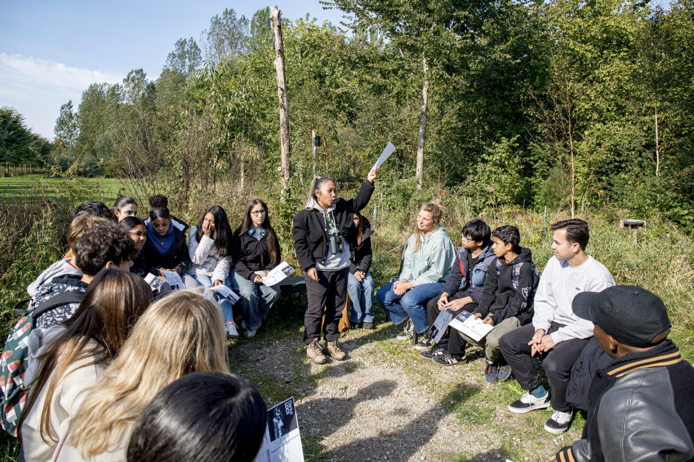 Een groep leerlingen zit in een halve cirkel in het Leeuwarder Bos. Een begeleider staat in het midden en houdt een papier omhoog terwijl ze iets uitlegt. De leerlingen luisteren aandachtig, met papieren of boekjes in hun handen. Op de achtergrond is groen struikgewas en een blauwe lucht te zien.