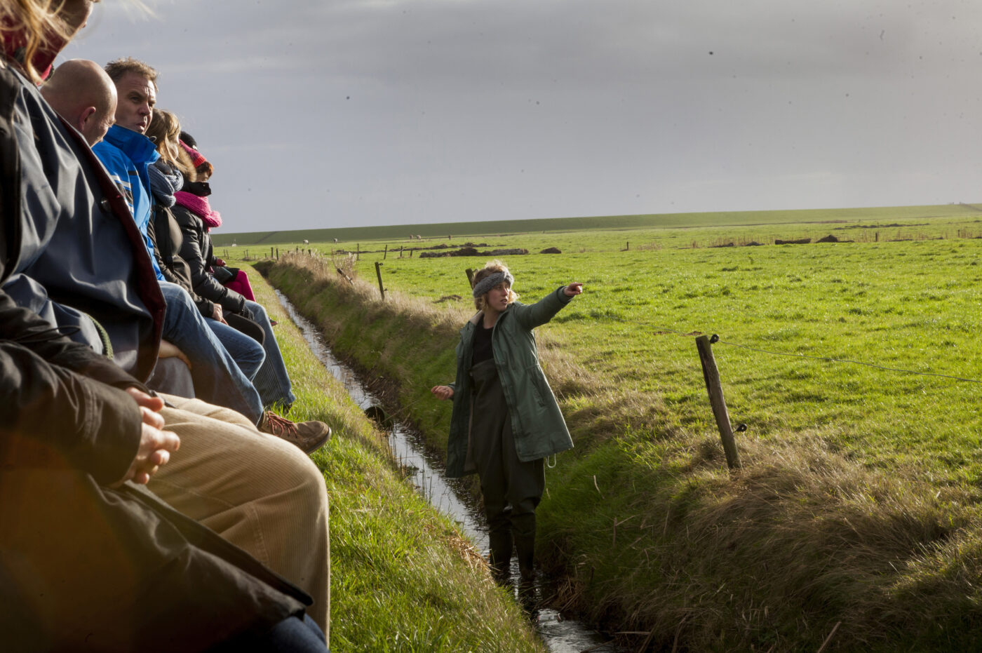 Een performer staat in een smal slootje tussen groene weilanden en wijst met gestrekte arm richting de horizon. Op de oever zit een groep mensen dicht naast elkaar, aandachtig toekijkend. Het zonlicht breekt door de wolken en verlicht het landschap.